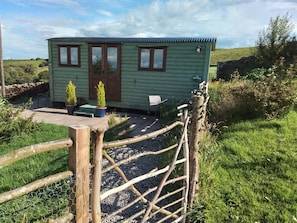 A small, green cottage with a wooden door, two windows, and a small porch area with a chair and a table. - The Herdsman, Barnoldswick (Barnoldswick)