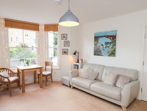 Ein gemütliches Wohnzimmer mit einem weißen Sofa, einem Holztisch und Stühlen. Es gibt ein großes Fenster mit Jalousien, eine Pendelleuchte und ein Bild an der Wand.