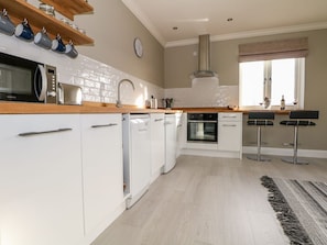 A modern kitchen with white cabinets, a microwave, oven, and a coffee machine. There are two bar stools and a window with a view. - Roughwood, Poulner, Hampshire (Ringwood)