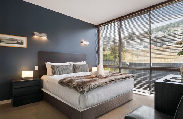 Une chambre à coucher moderne avec un grand lit, des murs de couleur sombre et une vue dégagée grâce à de grandes fenêtres du sol au plafond.