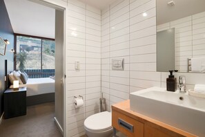 Une salle de bain moderne avec des carreaux blancs, des toilettes, un lavabo avec un miroir et une fenêtre avec des stores.