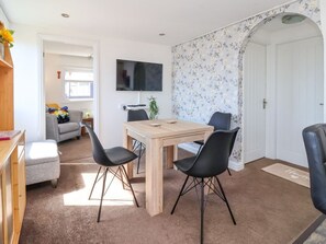 A room with a wooden dining table, four black chairs, a grey sofa, a flat-screen TV mounted on the wall, and a floral wallpaper pattern.