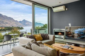 A modern living room with a large TV, a fireplace, and a scenic view of mountains and a lake. - 1-BR Pounamu Terrace | Central - Spacious (Queenstown)
