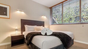 Quarto com cama, mesa de cabeceira, abajur, janela com persianas e um quadro na parede.