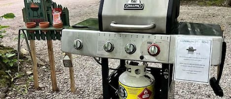 A portable grill with a propane tank, a 'COMMUNITY BBQ' sign, and a small table with a sign that reads 'It's BBQ Time.'