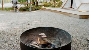 A fire pit with logs and a grill, a tent, and a person sitting on a bench in a wooded area.