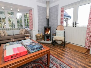 Ein gemütliches Wohnzimmer mit Kamin, einer Couch, einem Couchtisch mit Büchern und einem Fenster mit Vorhängen.