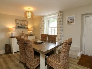 A dining room with a wooden table and wicker chairs, a window with curtains, and wall-mounted lamps. - Pheasant Cottage, Ilfracombe (Ilfracombe)