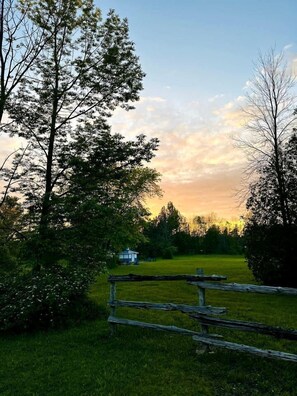 Un paysage rural avec une clôture en bois, un petit bâtiment blanc et des arbres sous un ciel au coucher du soleil.