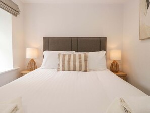 A neatly made bed with a headboard, two bedside lamps, and a window with curtains. - Riverside Cottage No 5 Keswick (Keswick)