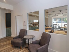 A modern interior with two armchairs and a small round table, a large mirror reflecting the room, and a view of a cafe area through the mirror. - Riverside Cottage No 5 Keswick (Keswick)