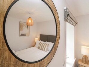 A bedroom with a bed, a round mirror, a hanging light fixture, a bedside table, and a window with curtains. - Riverside Cottage No 5 Keswick (Keswick)