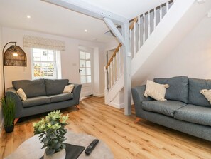 A living room with two grey sofas, a coffee table with a plant, a remote control, and a staircase leading to an upper floor. - Riverside Cottage No 5 Keswick (Keswick)