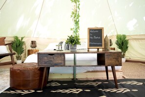 A tent interior with a bed, a wooden desk with drawers, a welcome sign, and potted plants.