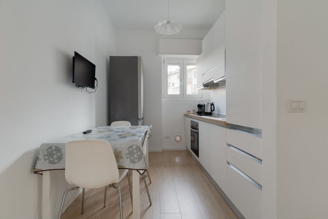 A small kitchen with a dining table, chairs, a TV mounted on the wall, and a window.