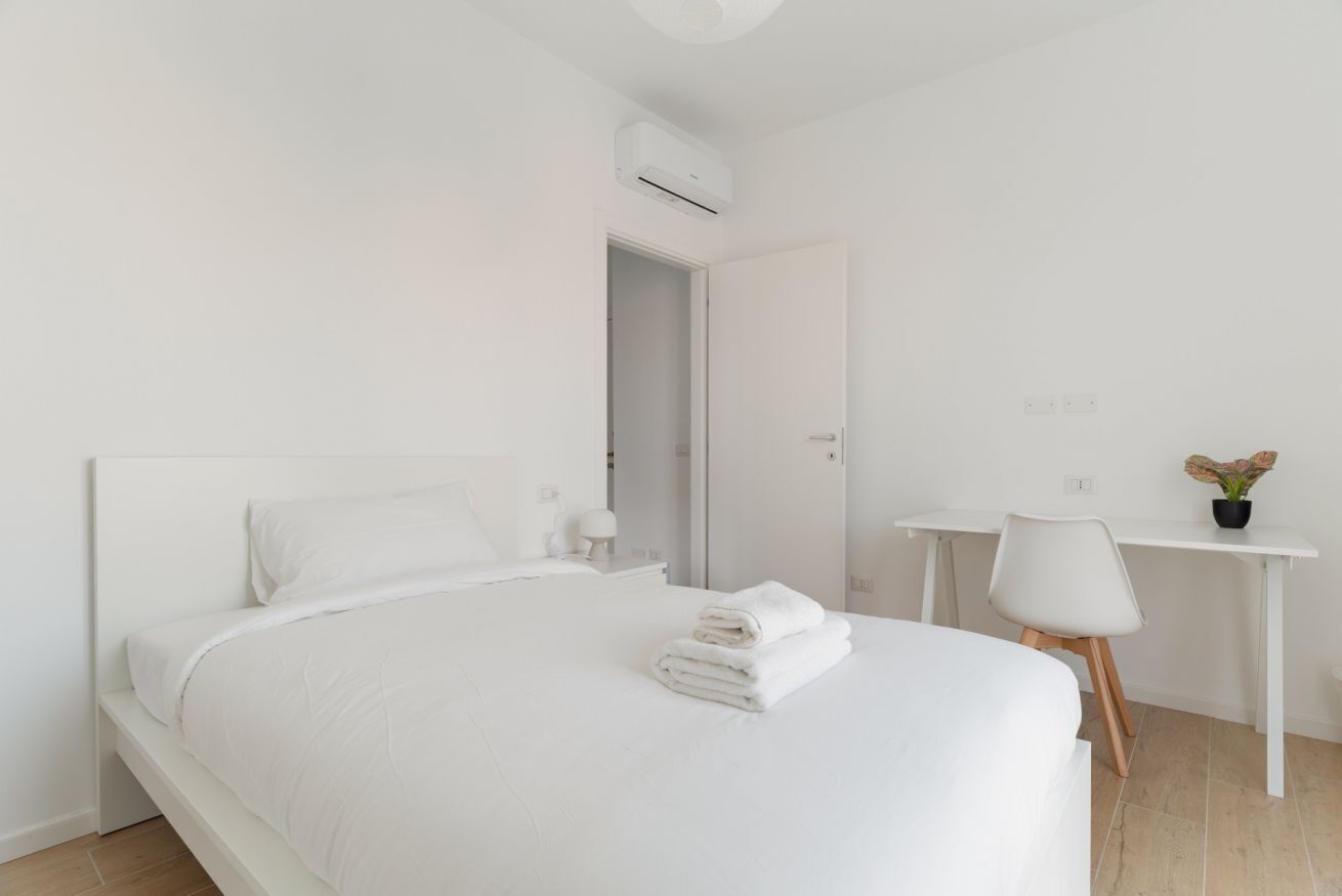 Een witte slaapkamer met een bed, nachtkastje, stoel, bureau en een airconditioning.