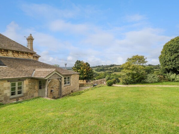 A stone house with a chimney, a grassy yard, and a view of rolling hills in the background.