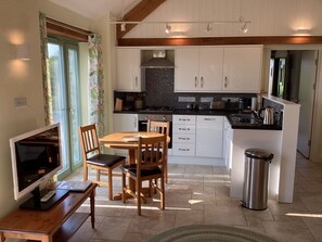A kitchen with white cabinets, a black countertop, a wooden dining table with chairs, a TV on a wooden stand, and a trash bin.
