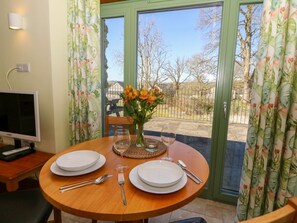 A dining table set with plates, cutlery, and a vase of flowers, with a view of trees and a fence through a glass door.