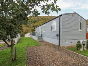 A single-story building with a gravel driveway, a small garden area with a tree, and a hill in the background.