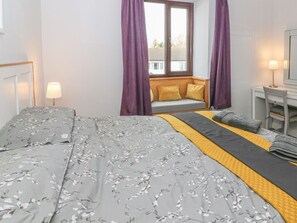 Ein Schlafzimmer mit einem Bett, einer Sitzbank am Fenster mit Kissen, einem Nachttisch mit Lampe und einem Spiegel.