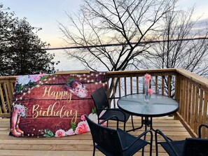 A wooden deck with a glass table and chairs, a 'Happy Birthday' sign, and a view of the water.