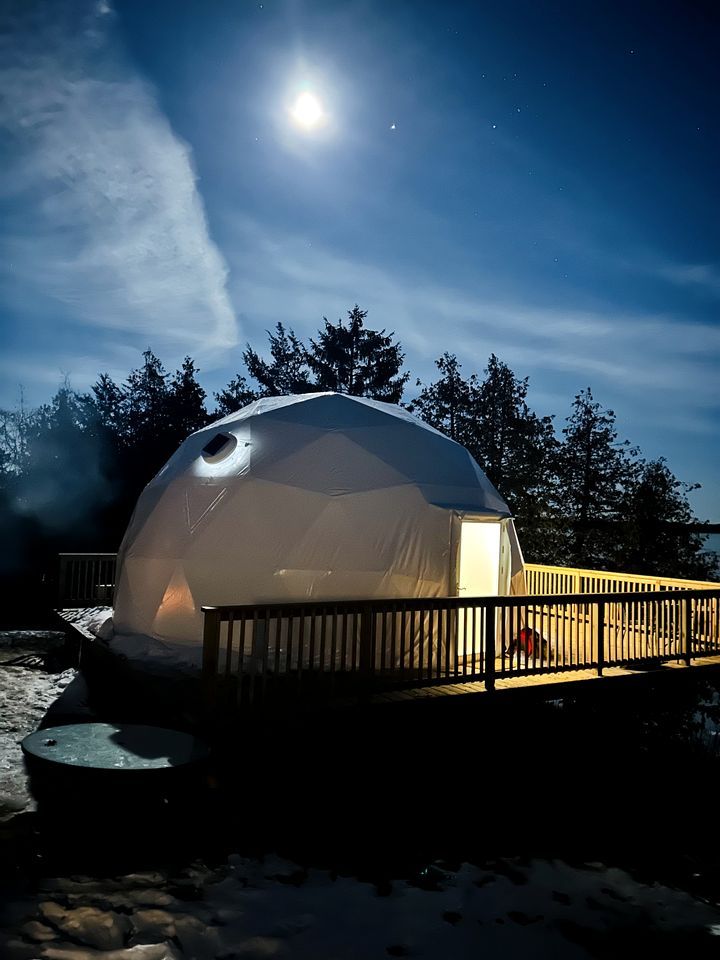 A dome-shaped tent with a wooden deck in front, set against a snowy landscape and a clear night sky.