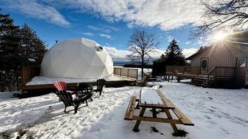 Une structure en forme de dôme géodésique, une terrasse en bois et une table de pique-nique, le tout dans un décor extérieur enneigé.