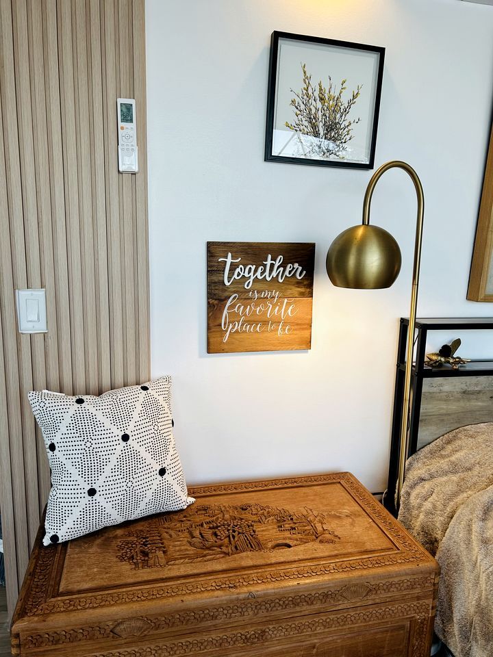Un dormitorio ordenado con un baúl de madera, una lámpara de pared, una cita inspiradora enmarcada y una pequeña fotografía enmarcada.