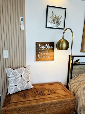 Un dormitorio ordenado con un baúl de madera, una lámpara de pared, una cita inspiradora enmarcada y una pequeña fotografía enmarcada.