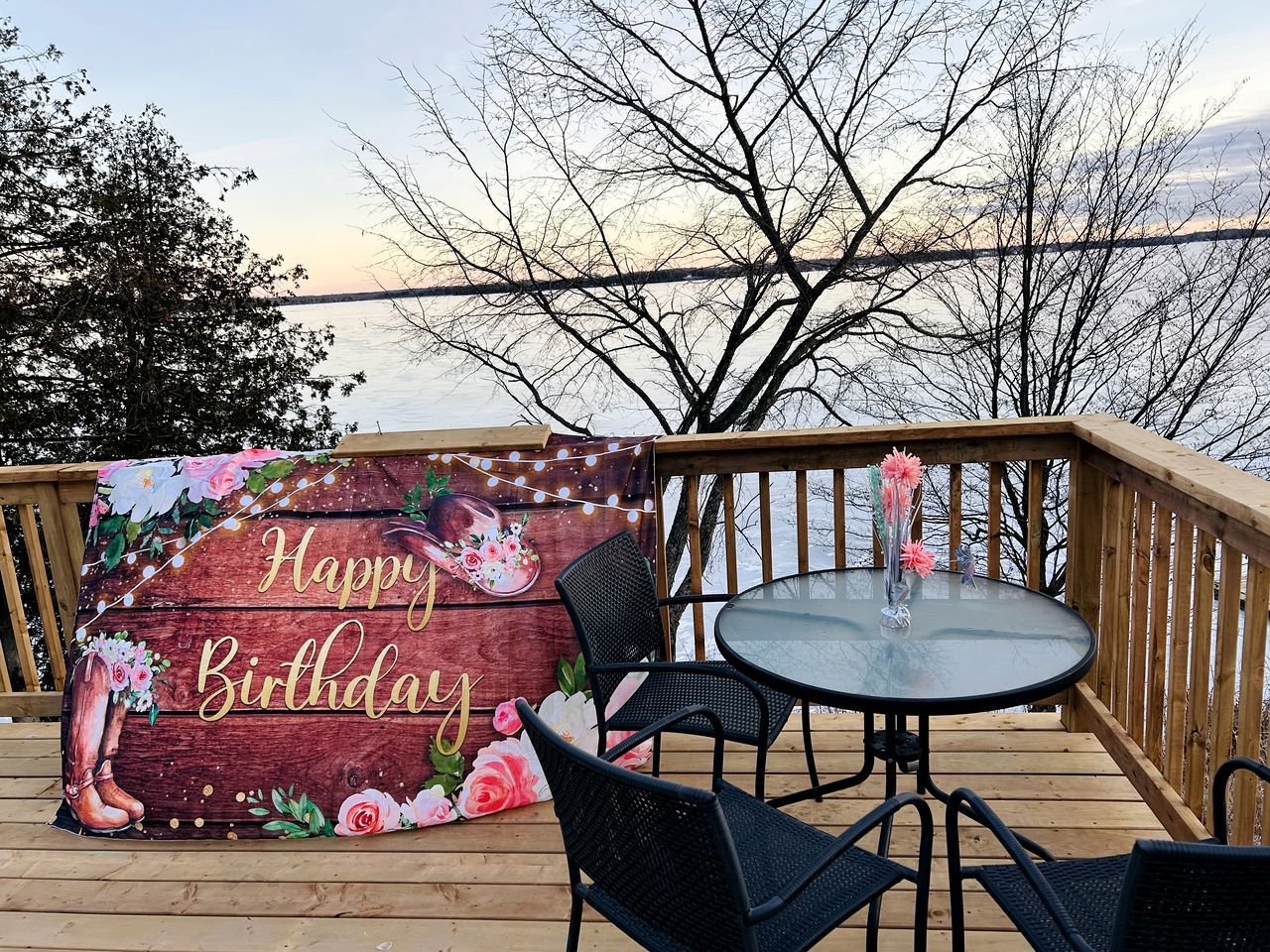 Um deck de madeira com mesa e cadeiras de vidro, um letreiro com a mensagem 'Happy Birthday' e vista para a água.