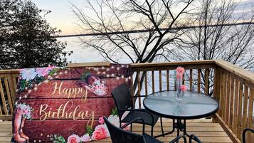 Um deck de madeira com mesa e cadeiras de vidro, um letreiro com a mensagem 'Happy Birthday' e vista para a água.