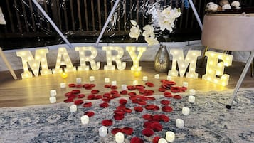 Une pancarte où est écrit « ÉPOUSE-MOI », avec des lettres illuminées, entourée de bougies et de pétales de roses rouges sur un tapis à motifs.