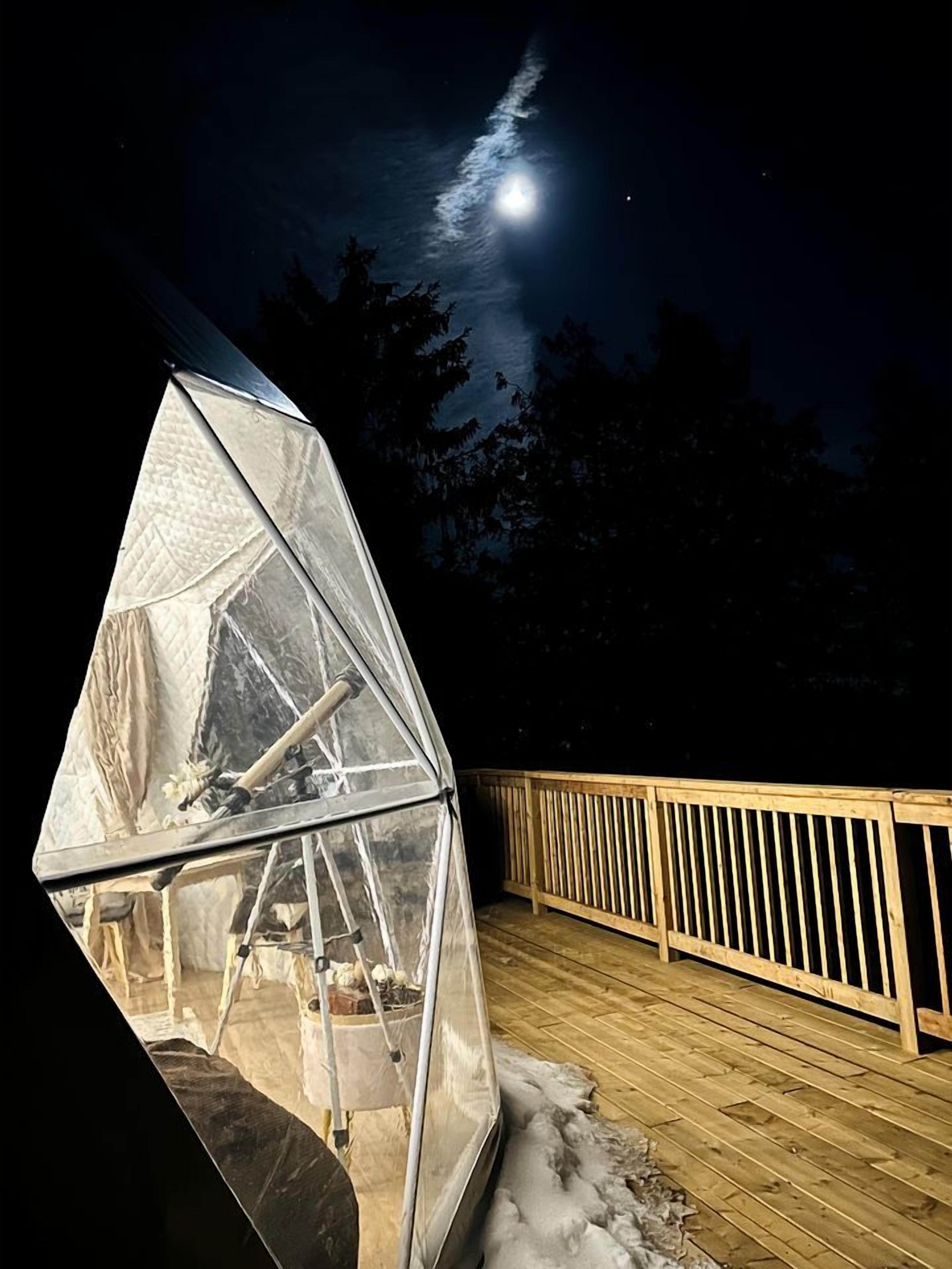 A transparent greenhouse structure on a wooden deck at night with a moon in the background.