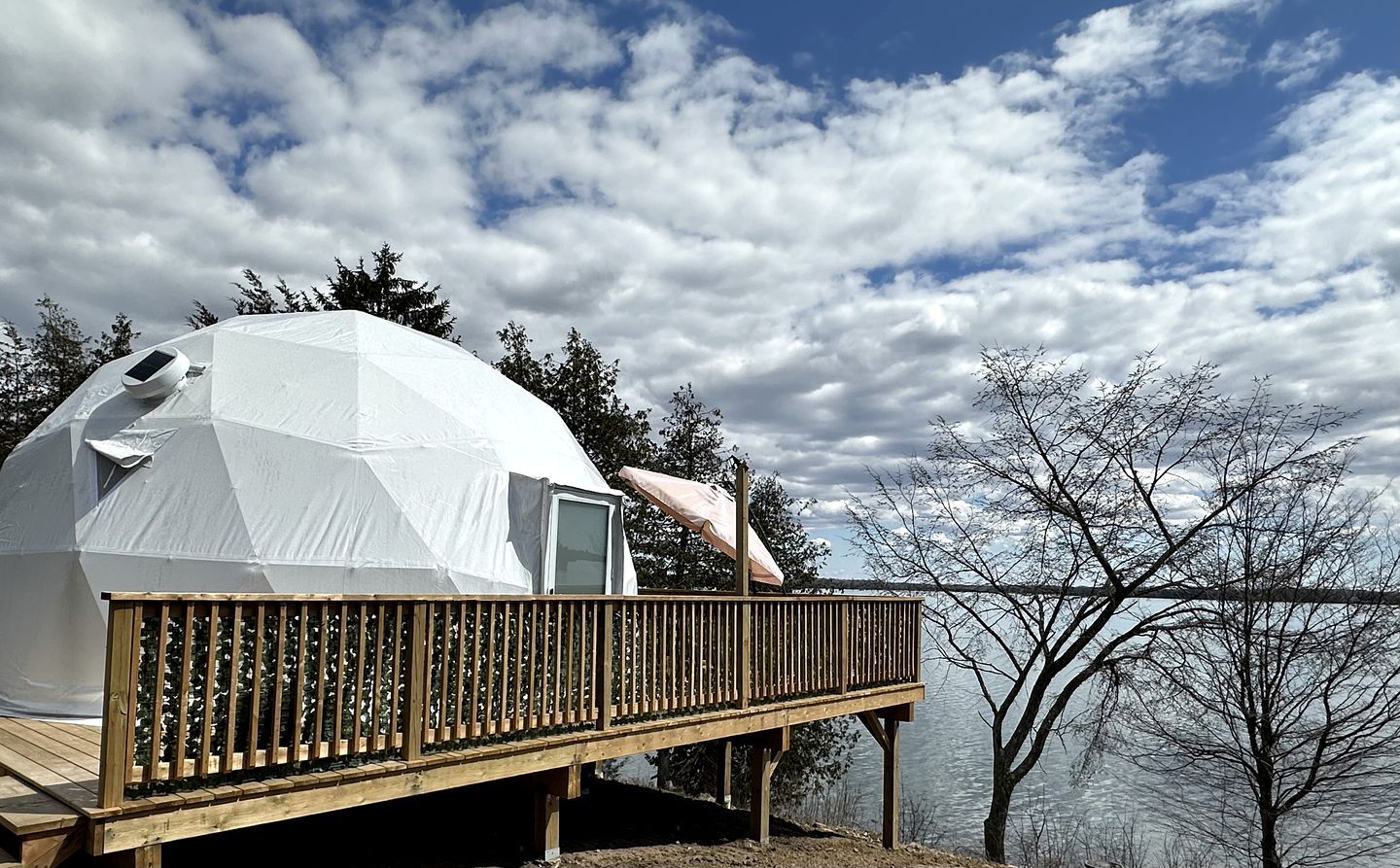 A geodesic dome structure with a wooden deck and a small enclosed area, overlooking a body of water and trees.