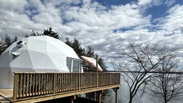 A geodesic dome structure with a wooden deck and a small enclosed area, overlooking a body of water and trees.