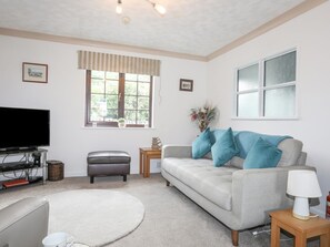 A living room with a flat-screen TV, a grey sofa with blue cushions, a small wooden side table, a white lamp, and a window with a view of greenery.