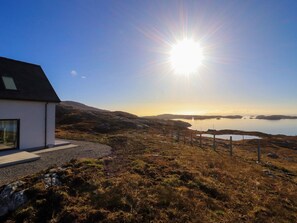 A modern house with a large window, a gravel driveway, and a view of a body of water and hills. - 8B Carrigreich, Tarbert Isle Of Harris (Isle of Harris)