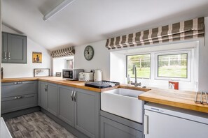 A modern kitchen with grey cabinets, a wooden countertop, a white sink, a clock, and a window with a view of greenery. - Prince's Cottage, Moniaive (Thornhill)