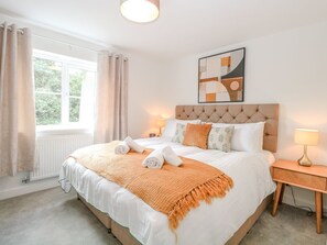 Ein ordentlich eingerichtetes Schlafzimmer mit einem großen Bett, einem Nachttisch, einem Fenster mit Vorhängen und einem an der Wand hängenden abstrakten Gemälde.