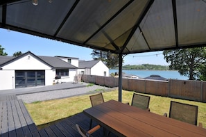 Un espacio de patio cubierto con una mesa y sillas, una parrilla de barbacoa y vista a un lago y casas al fondo.