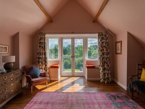 A room with a sloped ceiling, wooden beams, a large window with a view of greenery, a bench with cushions, a dresser, and a rug. - Coach House, Llanwrtyd Wells (United Kingdom)