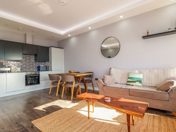 A modern kitchen with dark cabinets, a dining area with a wooden table and chairs, a sofa with striped cushions, and a round mirror on the wall.