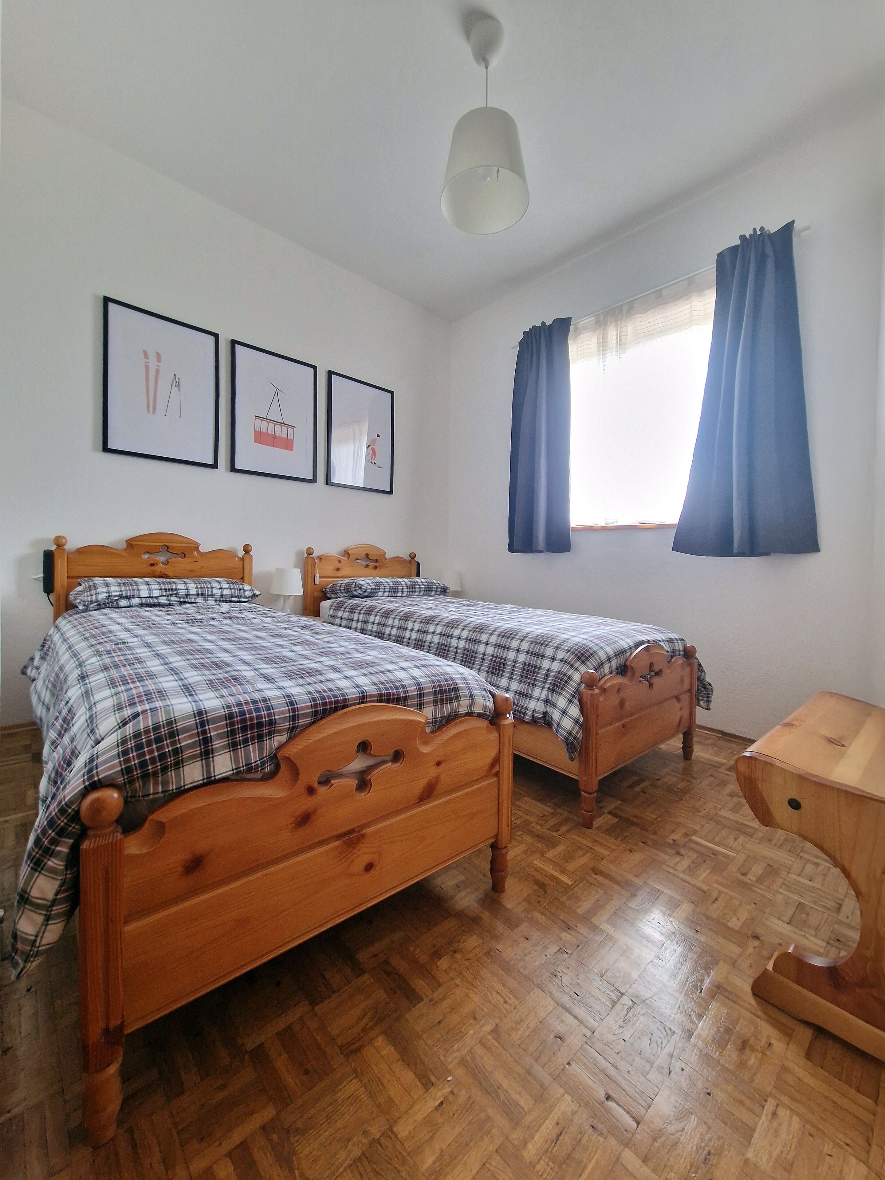 A room with two wooden beds, a ceiling light, and three framed pictures on the wall.