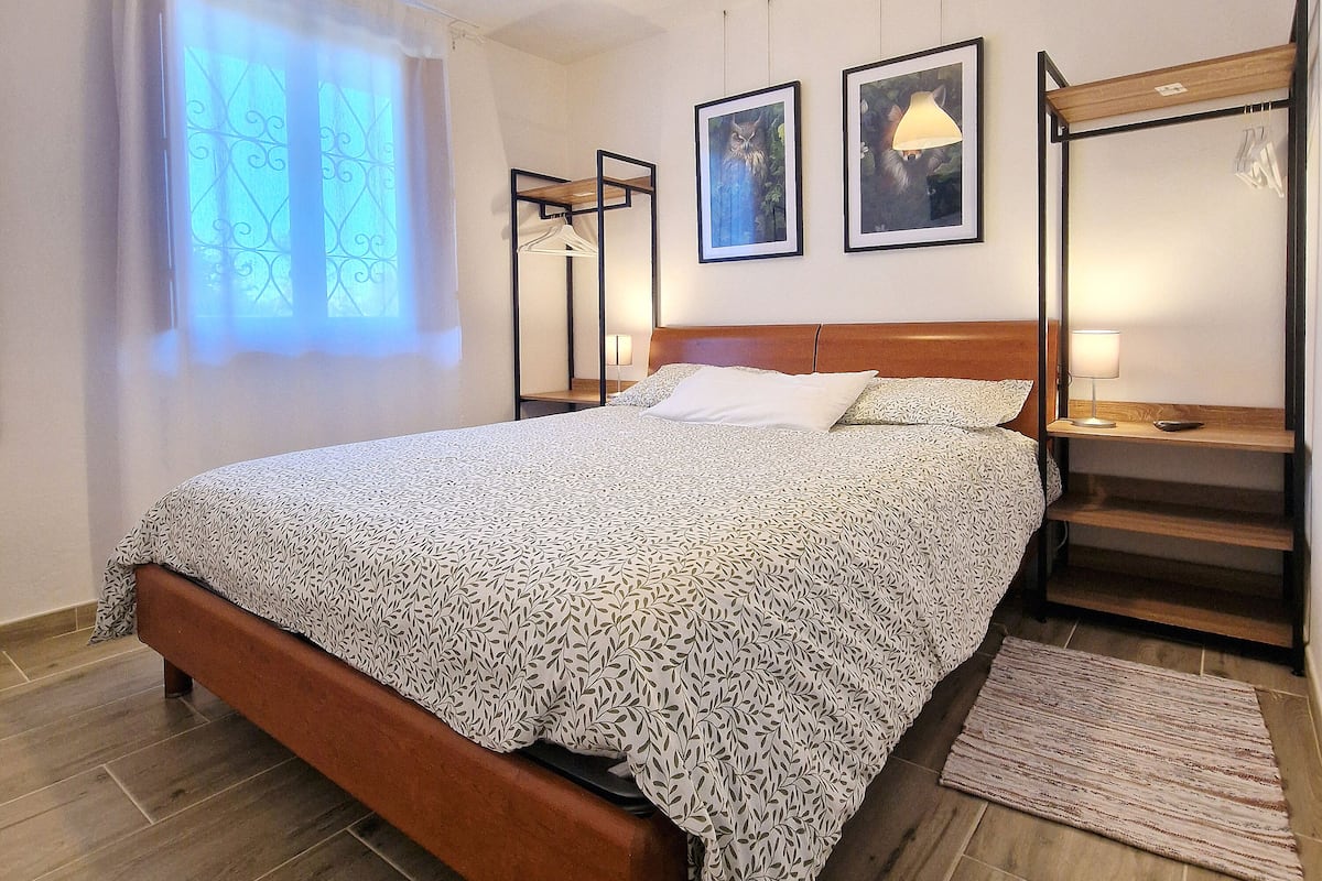 A bedroom with a bed, a wooden headboard, a nightstand, a lamp, a framed picture, a window with curtains, and a wooden floor.