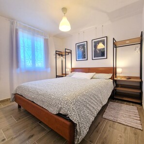 A bedroom with a bed, a wooden headboard, a nightstand, a lamp, a framed picture, a window with curtains, and a wooden floor. - Casa Polsa 1964 (Polsa)