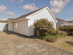 A single-story building with a sloped roof, a small porch, and a garden with flowering plants. - Seaside retreat, Porthmadog (Porthmadog)