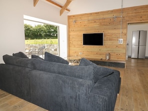 Ein modernes Wohnzimmer mit einem großen grauen Sofa, einem flachbildfernseher an einer Holzwand und einer Küchenzeile mit einem Edelstahlkühlschrank.