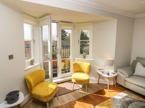 A living room with a yellow armchair, a sofa, a small table, and a window with a view of a balcony. - Seashells, Tenby, Pembrokeshire (Tenby)