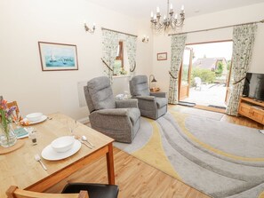 Ein Wohnzimmer mit zwei grauen Sesseln, einem Holzespeisetisch für zwei Personen, einer TV-Kommode und einem großen Fenster mit Vorhängen.